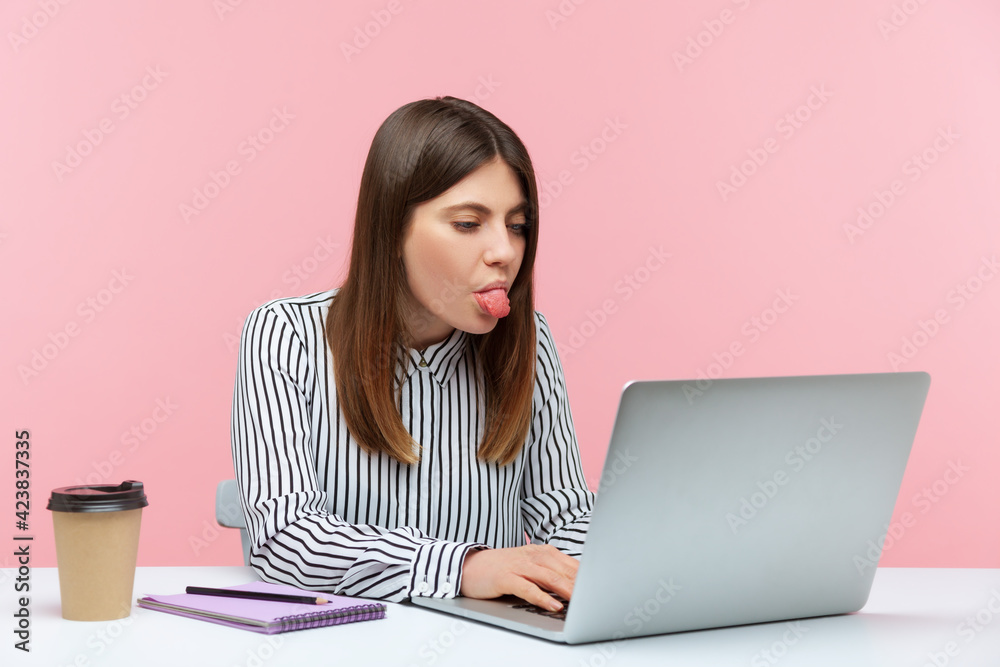 Naklejka premium Playful funny woman office worker in striped shirt showing tongue out, expressing disgust and disobedience chatting via web camera on laptop. Indoor studio shot isolated on pink background