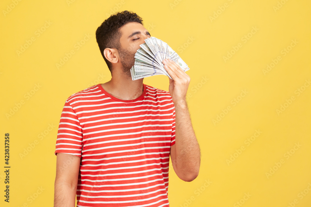 Greedy mercantile man in red striped t-shirt smelling fan of cash, enjoying aroma of hundred dollars bills in his hand, satisfied with big profit. Indoor studio shot isolated on yellow background Greedy mercantile man in red striped t-shirt smelling fan of cash, enjoying aroma of hundred dollars bills in his hand, satisfied with big profit. Indoor studio shot isolated on yellow background