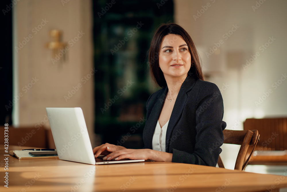 The assistant works on a laptop computer in the office. Online foreign language training.Portrait of a beautiful young woman sitting at a wooden table. Comfortable working space in a coworking space. The assistant works on a laptop computer in the office. Online foreign language training.Portrait of a beautiful young woman sitting at a wooden table. Comfortable working space in a coworking space.