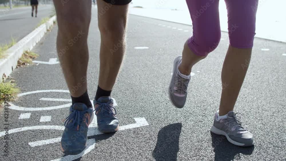 Male and female legs running on asphalt road. Unrecognizable joggers or runners jogging together outdoors in sunny day. Closeup view. Tracking camera. Healthy lifestyle and sport activity concept Male and female legs running on asphalt road. Unrecognizable joggers or runners jogging together outdoors in sunny day. Closeup view. Tracking camera. Healthy lifestyle and sport activity concept