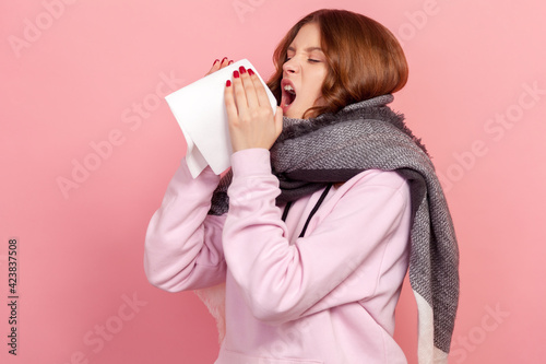 Side view portrait of ill curly haired teenage girl in hoodie and warm scarf coughing, sneezing hard in napkin, feeling unwell with runny nose, allergy. Indoor studio shot isolated on pink background