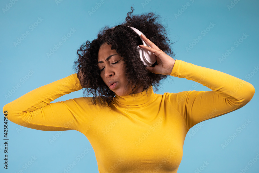 African woman dancing with wireless headphones isolated on blue studio wall. Cute lady in yellow wear, she is singing. Music, radio, happiness, freedom, youth concept.