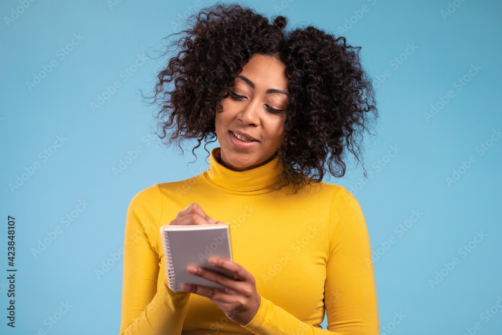Obraz premium African woman making notes in planner, mature lady holding pen. She writes future plans and to-do list in notebook for week or month. Keeping personal diary on blue studio background.
