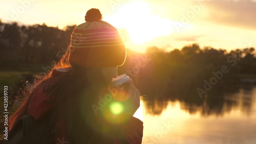 Woman Traveler holds mug with hot coffee in her hands and looks at sunset byriver. A tourist drinks tea from a mug in sun. Free traveler girl admiring scenery. Hiking. Hiker drinking hot cocoa