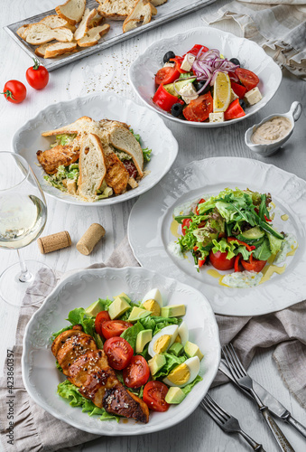 Fresh salads with grilled chicken meat, vegetables, eggs, greens, feta cheese, olives, parmesan, lettuce, tomatoes on plates over light wooden background. Healthy food, clean eating, top view