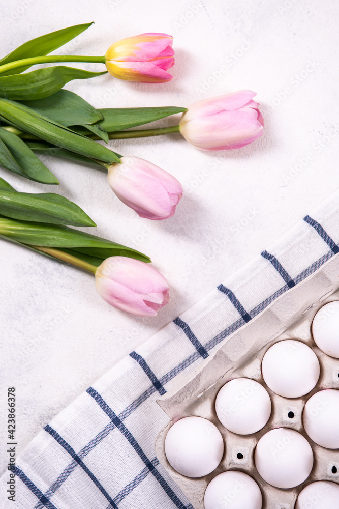 White chicken eggs in the tray, on a blue and white checkered napkin. Nearby are pink tulips. Top view, flat lay.
