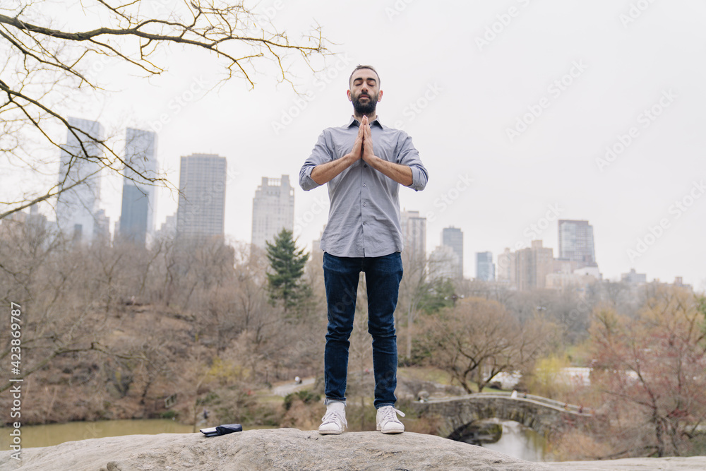 Man with beard meditates in city the park nature, standing. Wearing formal shirt and casual jeans. Namaste praying hands in front of the heart Man with beard meditates in city the park nature, standing. Wearing formal shirt and casual jeans. Namaste praying hands in front of the heart