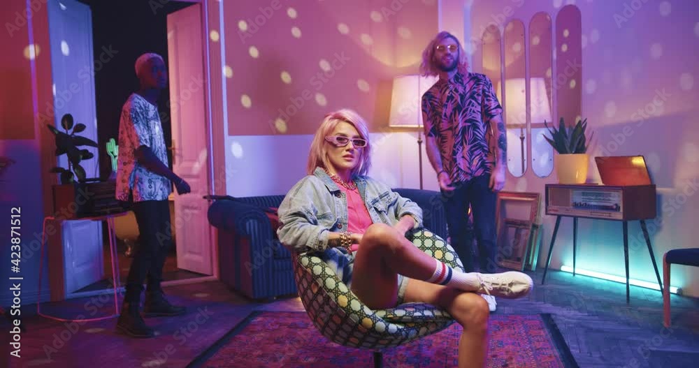 Three multinational hipster friends had retro-style home party. Stylish girl sitting on chair on background of dancing men.