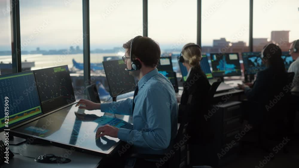Male Air Traffic Controller with Headset Using Touchscreen Table Panel in Airport Tower. Office Room is Full of Computers with Navigation Screens, Airplane Departure and Arrival Data for the Team. Male Air Traffic Controller with Headset Using Touchscreen Table Panel in Airport Tower. Office Room is Full of Computers with Navigation Screens, Airplane Departure and Arrival Data for the Team.