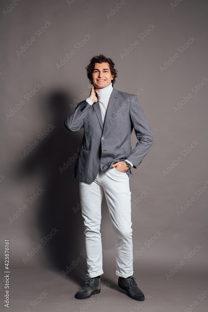 Tall handsome man dressed in white turtleneck, white jeans and grey jacket posing on the grey background Tall handsome man dressed in white turtleneck, white jeans and grey jacket posing on the grey background