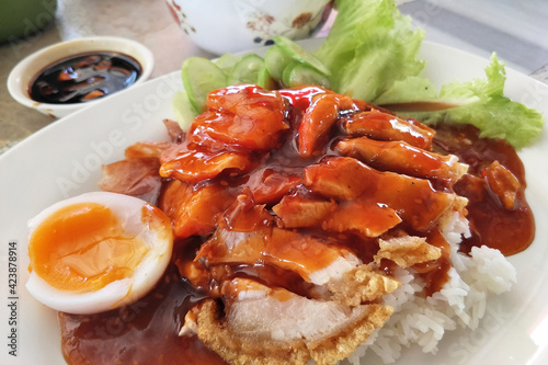 Rice with red barbecue pork, Crispy Pork, and Egg topped with sauce in a white dish. It is a delicious menu of Thai.