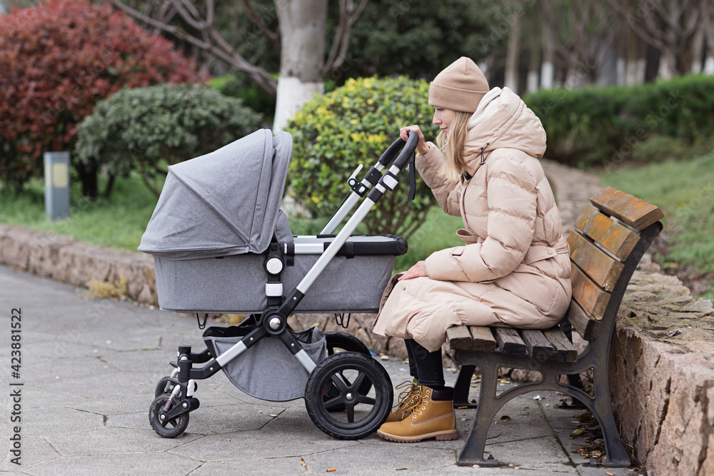 Happy Mother walking With Stroller In Park. Joy of motherhood. Stylish young caucasian woman wearing warm clothes at autumn.