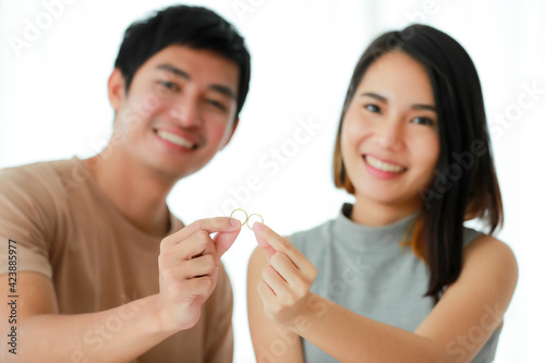 Front view portrait of cute smiling young Asian lover couple in casual clothes, looking at the camera and showing a golden engagement rings together. Selective focus at the rings with a blurred face
