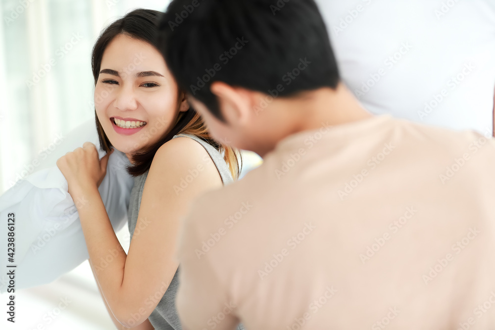 Funny shot of cute smiling young adult Asian woman in casual clothes having fun with a pillow fight with her husband in the foreground. Wife and husband playing spend time together in the morning Funny shot of cute smiling young adult Asian woman in casual clothes having fun with a pillow fight with her husband in the foreground. Wife and husband playing spend time together in the morning