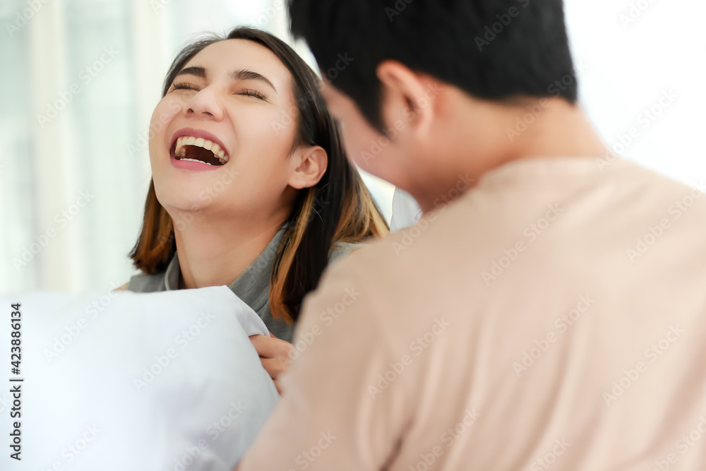 Funny shot of cute smiling young adult Asian woman in casual clothes having fun with a pillow fight with her husband in the foreground. Wife and husband playing spend time together in the morning