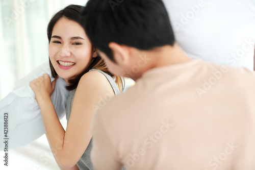 Funny shot of cute smiling young adult Asian woman in casual clothes having fun with a pillow fight with her husband in the foreground. Wife and husband playing spend time together in the morning
