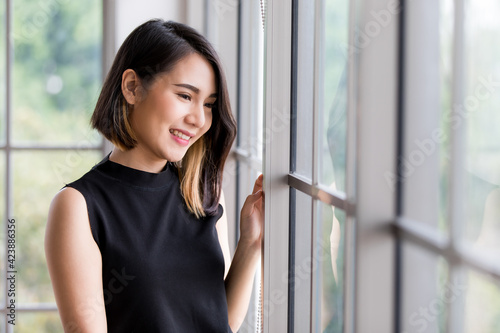 Horizontal portrait shot of a cute smiling Asian woman with a stylish straight hairstyle in black clothes standing, touching the windows, and looking at a tree outside. Concept of happiness at home
