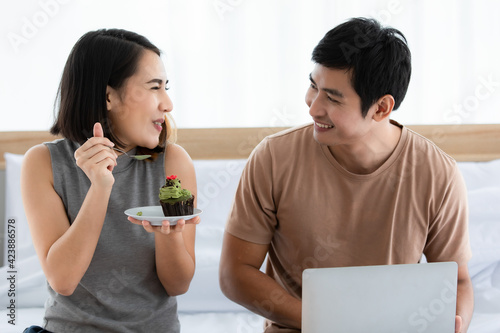 Portrait shot of cute smiling young Asian lover couple sitting on a bed at home and eating cake together. Love monent and sweet time sharing concept. photo