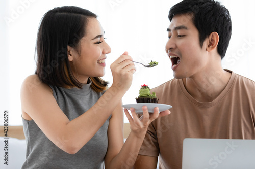 Portrait shot of cute smiling young Asian lover couple sitting on a bed at home and eating cake together. Love monent and sweet time sharing concept. photo