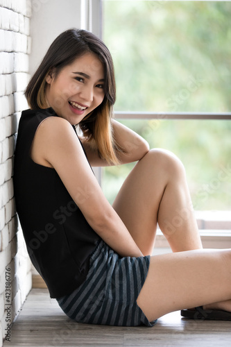 Vertical portrait shot of a cute smiling Asian woman with a straight hairstyle in black clothes sitting on the floor and leaning against the wall .near a window and looking at the camera in the studio