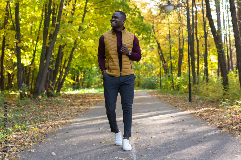 Positive african american young man in stylish clothes walks in late summer park on sunny warm warm day. Outdoor activity concept. Positive african american young man in stylish clothes walks in late summer park on sunny warm warm day. Outdoor activity concept.