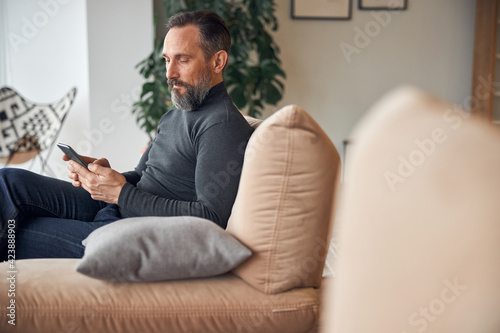 Caucasian adult man is sitting with smartpone in modern cozy office lounge zone