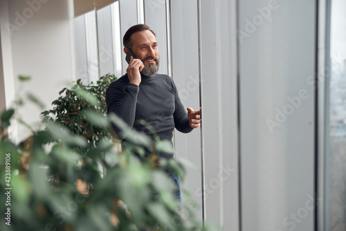 Happy smart adult caucasian businessman is standing near big windows in office space and holding smartpone