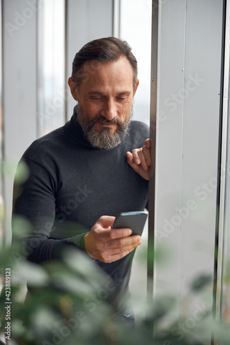 Happy smart adult caucasian businessman is standing near big windows in office space and holding smartpone