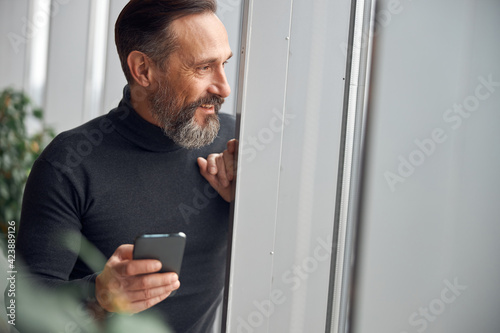 Happy smart adult caucasian businessman is standing near big windows in office space and holding smartpone