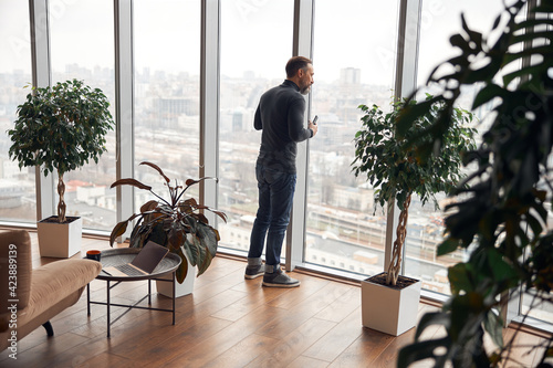 Happy smart adult caucasian businessman is standing near big windows in office space and holding smartpone