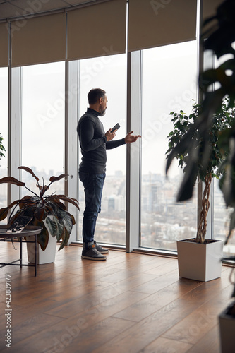 Happy smart adult caucasian businessman is standing near big windows in office space and holding smartpone