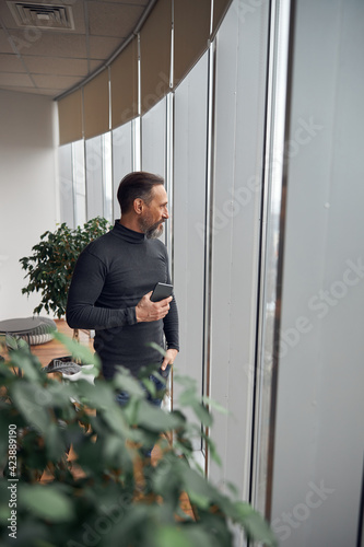 Happy smart adult caucasian businessman is standing near big windows in office space and holding smartpone