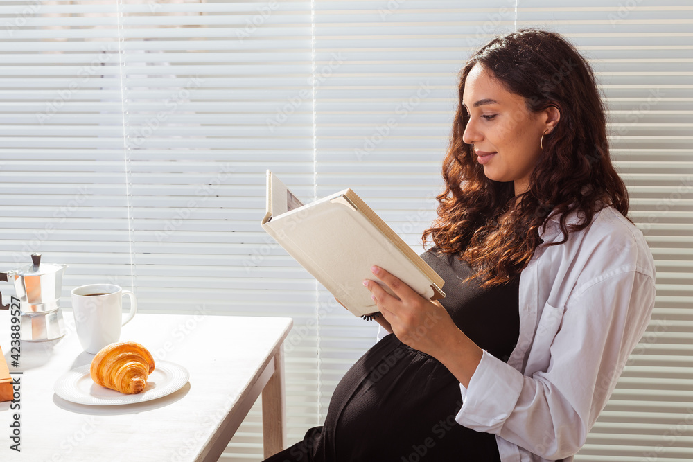 Side view of pregnant woman reading book while having morning breakfast with coffee and croissants on background of blinds. Good morning concept and pleasant lunch break Side view of pregnant woman reading book while having morning breakfast with coffee and croissants on background of blinds. Good morning concept and pleasant lunch break