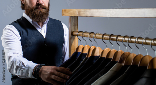 Tailor, tailoring. Stylish men's suit. Handsome bearded fashion man in classical costume suit. Man suit, tailor in his workshop. Male suits hanging in a row photo