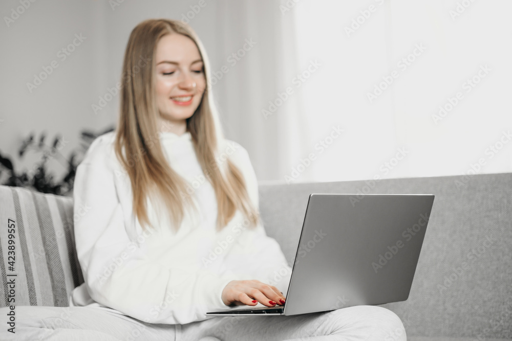 Young blonde woman sitting at home on the couch with laptop smiles and works distance in quarantine during the coronavirus pandemic Young blonde woman sitting at home on the couch with laptop smiles and works distance in quarantine during the coronavirus pandemic