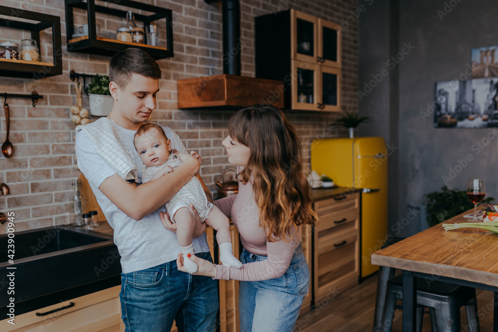 Fototapeta premium Young father holding baby boy in arms. Daddy and son together at home. Happy family in the kitchen. Mother looks at baby.