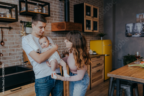Young father holding baby boy in arms. Daddy and son together at home. Happy family in the kitchen. Mother looks at baby.