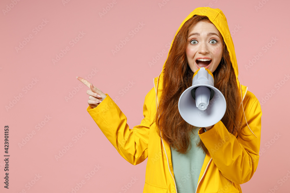 Redhead young woman 20s in yellow waterproof hood raincoat outerwear scream shout in megaphone point index finger aside on workspace isolated on pink background studio. Outdoors fall season concept Redhead young woman 20s in yellow waterproof hood raincoat outerwear scream shout in megaphone point index finger aside on workspace isolated on pink background studio. Outdoors fall season concept
