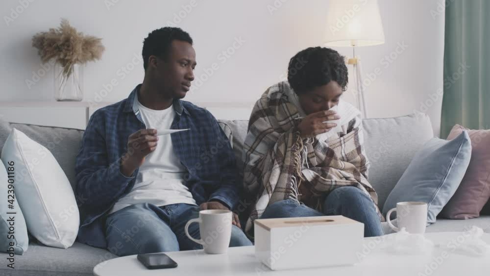 Serious disease. Black guy looking at thermometer of his sick wife, woman suffering from cough attack at home
