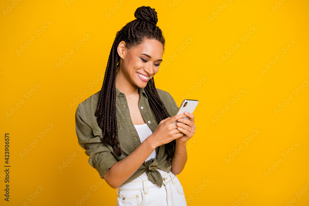 Photo of cheerful pretty dark skin blogger look phone write post comment isolated on yellow color background Photo of cheerful pretty dark skin blogger look phone write post comment isolated on yellow color background