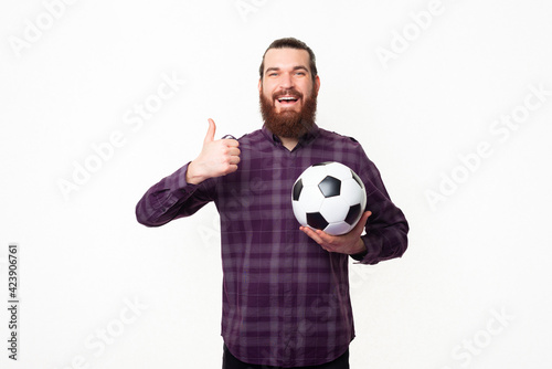 Photo of young bearded man showing thumb up and holding soccer ball