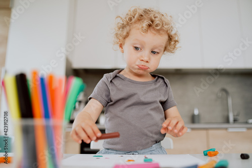 Portrait of a lovely happy preschool-aged girl sitting at her kitchen table enjoying some free time. Little cute girl is making something out of plasticine. She is making a funny face