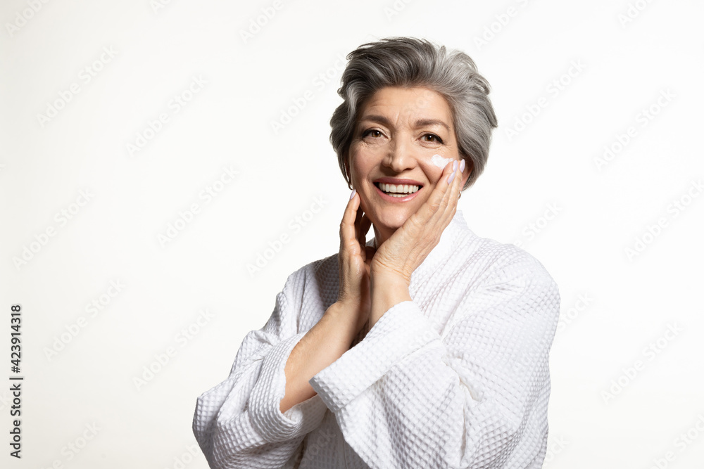 Senior female in bathrobe applying moisturizing creme on face looking at camera. Gray-haired laughing mature woman doing daily beauty routine. Elderly health, skincare and cosmetology Senior female in bathrobe applying moisturizing creme on face looking at camera. Gray-haired laughing mature woman doing daily beauty routine. Elderly health, skincare and cosmetology