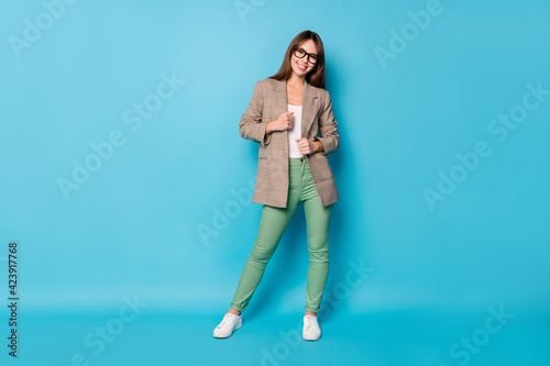 Full size photo of optimistic brunette lady stand wear spectacles brown jacket pants sneakers isolated on vivid blue background