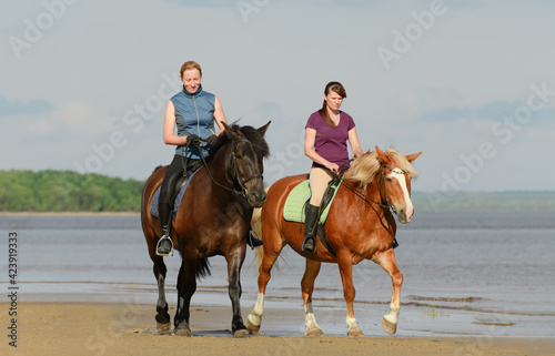 Two Caucasian women are riding on horseback on beach near a water.