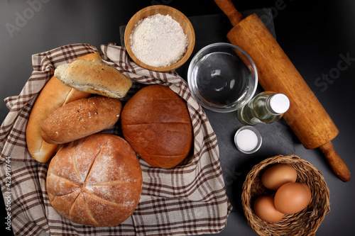 Freshly baked bread loaf bun roll round long mix verity wrapped in checkered kitchen fabric napkin towel wheat flower oil water salt eggs rolling pin slate stone over black background photo