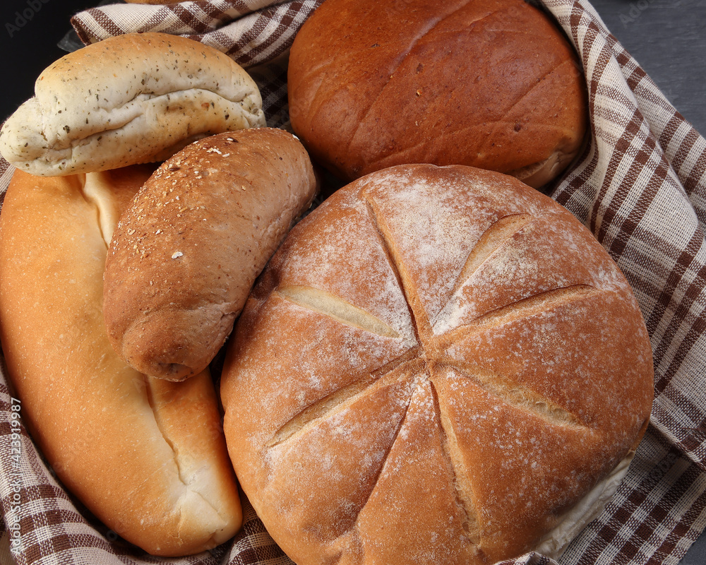 Freshly baked bread loaf bun roll round long mix verity wrapped in checkered kitchen fabric napkin towel wheat flower oil water salt eggs rolling pin slate stone over black background Freshly baked bread loaf bun roll round long mix verity wrapped in checkered kitchen fabric napkin towel wheat flower oil water salt eggs rolling pin slate stone over black background