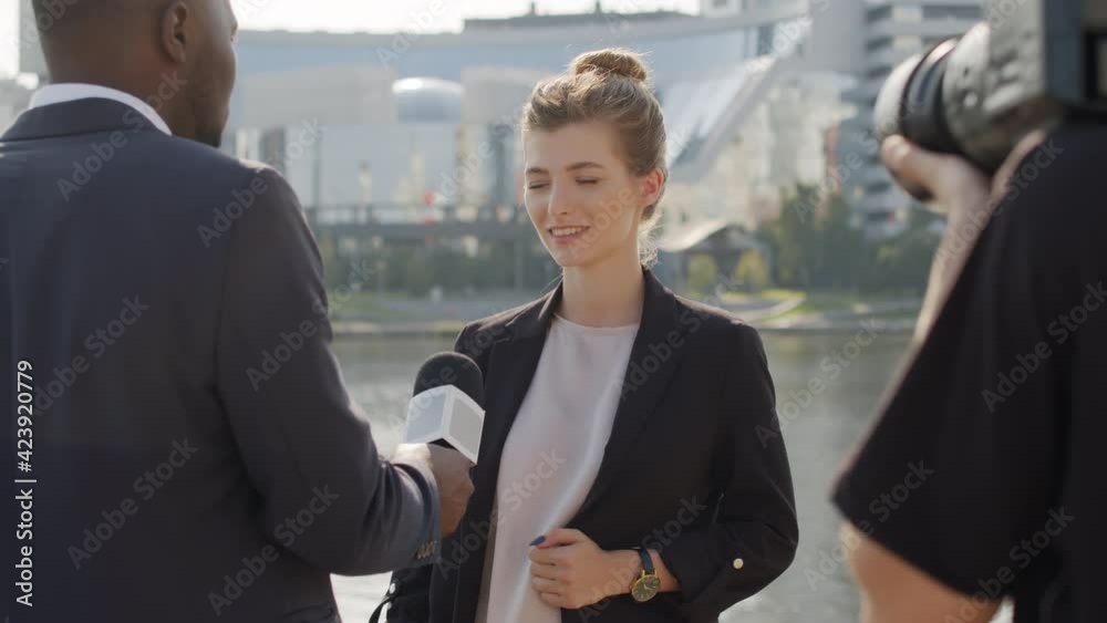 Medium shot of attractive young woman being interviewed by afro-american journalist outside in city centre while operator with camera filming them Medium shot of attractive young woman being interviewed by afro-american journalist outside in city centre while operator with camera filming them