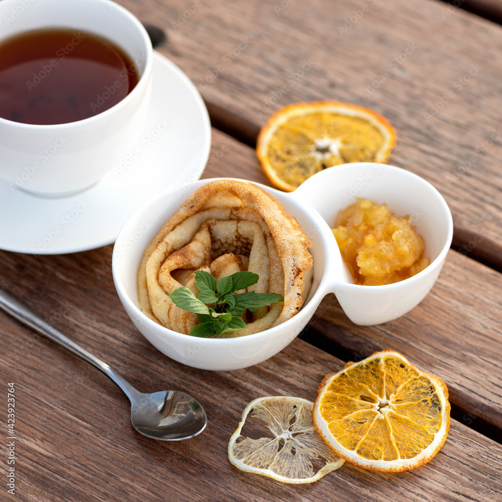 Tasty breakfast. Simple crepe, tea and citrus jam. Delicate thin pancake rolled on plate and saucer with orange jam. Wooden table on background. Top view. Good morning concept. Tasty breakfast. Simple crepe, tea and citrus jam. Delicate thin pancake rolled on plate and saucer with orange jam. Wooden table on background. Top view. Good morning concept.