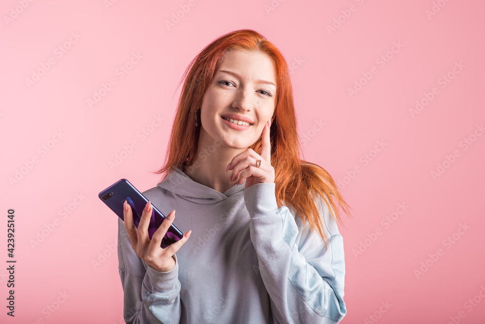 Portrait of a pensive redhead girl with a smartphone in her hands in the studio on a pink background Portrait of a pensive redhead girl with a smartphone in her hands in the studio on a pink background